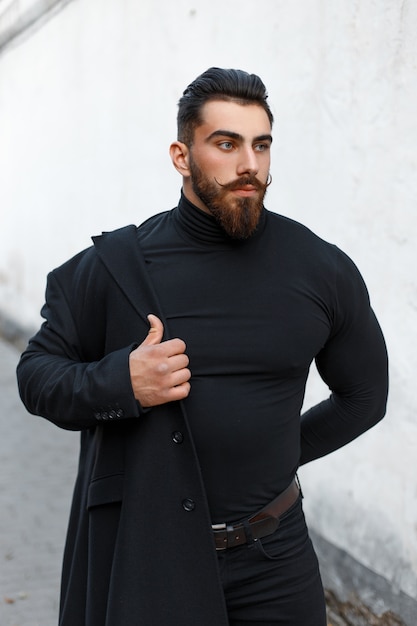 Hombre guapo de moda con peinado y barba se pone un elegante abrigo negro en la calle