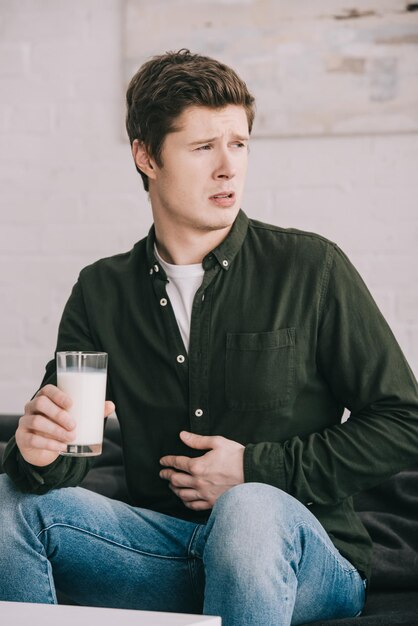 Hombre guapo y molesto con un vaso de leche sosteniendo el estómago