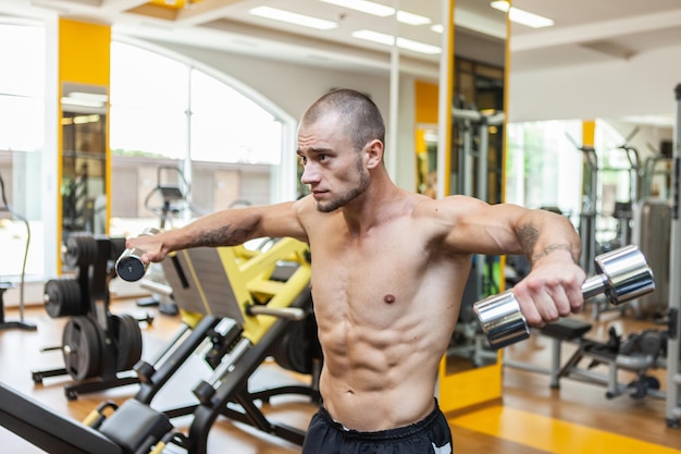 Hombre guapo musculoso culturista entrena los músculos del hombro levantando pesas con las manos a los lados en el gimnasio