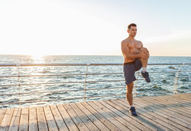 Hombre guapo musculoso con el torso desnudo haciendo ejercicio de calentamiento antes de hacer ejercicio en la playa al amanecer.