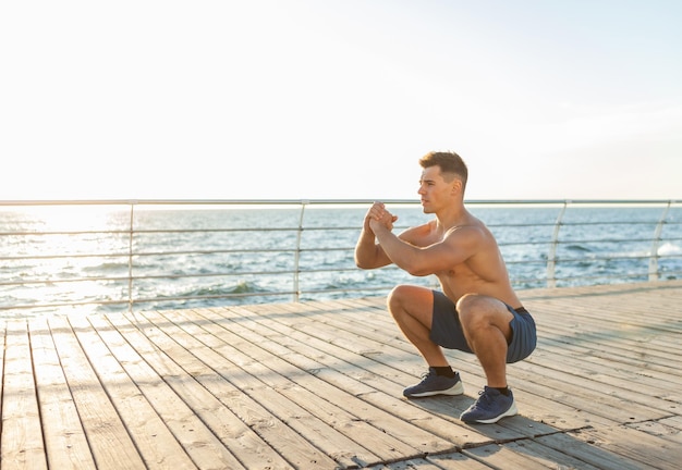 Hombre guapo musculoso con el torso desnudo haciendo ejercicios en cuclillas en la playa al amanecer.