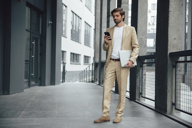 Hombre guapo ocupado en traje formal de moda sosteniendo en una mano moderno smartphone y portátil en otra. Empresario barbudo de pie cerca del centro de la oficina