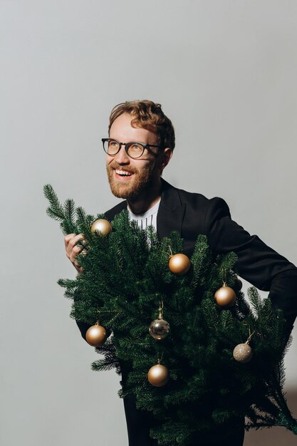 Hombre guapo pelirrojo con una chaqueta negra y gafas baila con un árbol de Navidad en sus manos Concepto de fiesta de Año Nuevo