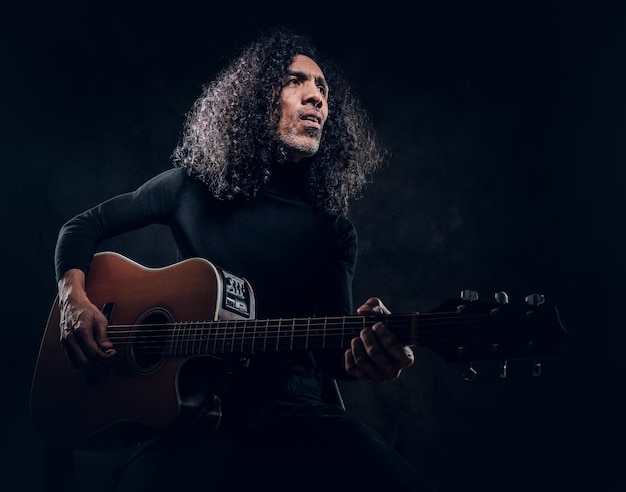 Un hombre guapo con el pelo largo toca la guitarra acústica en un estudio oscuro.