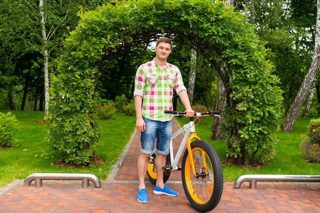 Hombre guapo de pie en la acera delante del arco de arbustos con bicicleta en un parque