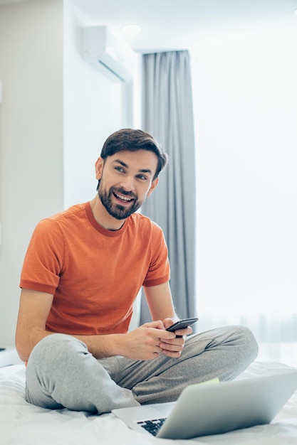 Hombre guapo positivo con ropa informal sentado en la cama frente a una computadora portátil y mirando hacia otro lado con una sonrisa mientras sostiene un teléfono inteligente