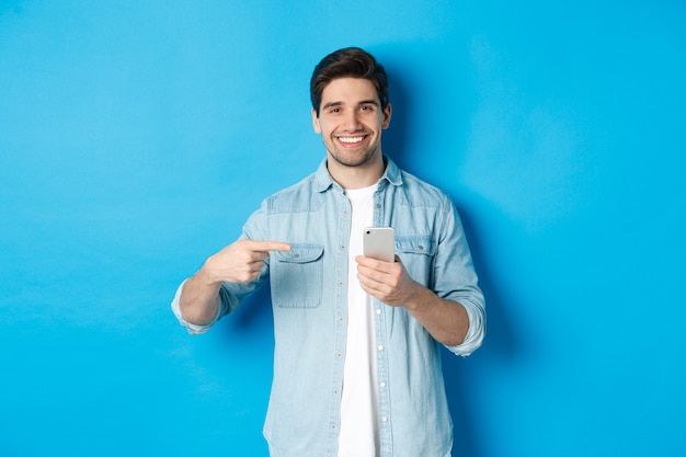 Hombre guapo recomendando la aplicación en el teléfono inteligente, apuntando al teléfono y sonriendo satisfecho, de pie sobre la pared azul