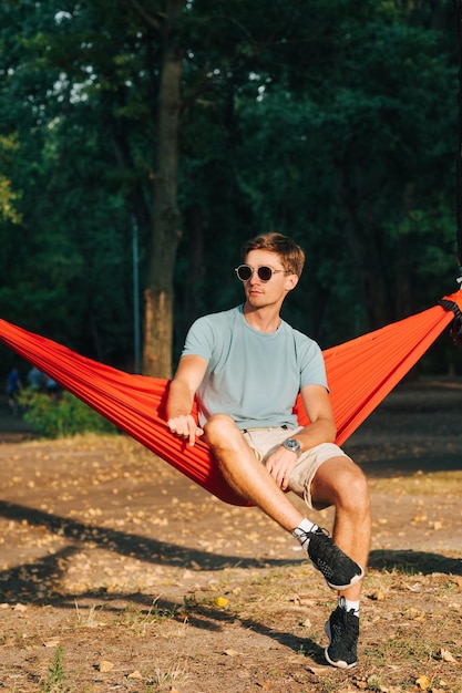 hombre guapo con ropa ligera e informal descansando sentado en una hamaca en el parque al atardecer