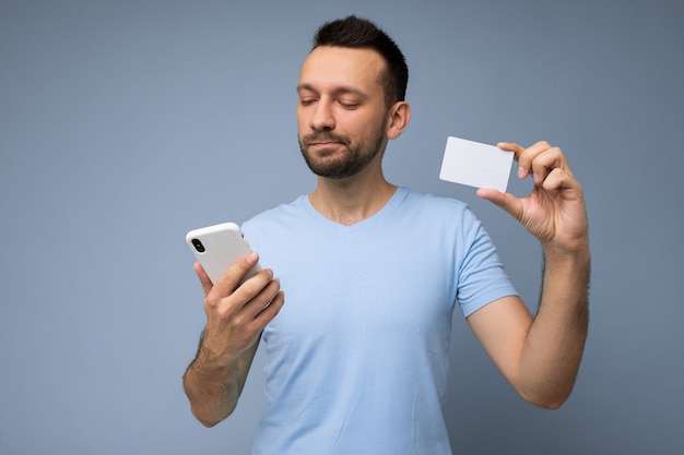 Hombre guapo con ropa de todos los días aislado en la pared de fondo sosteniendo y usando teléfono y crédito