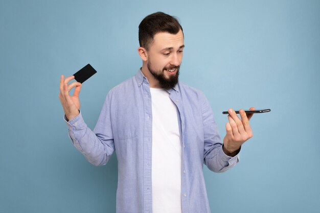 Hombre guapo con ropa de todos los días aislado en la pared de fondo sosteniendo y usando teléfono y crédito