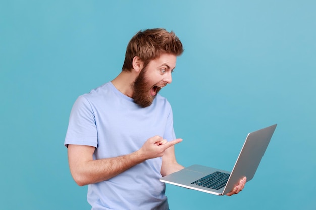 Hombre guapo señalando con el dedo la pantalla del portátil con grandes ojos impactados con el contenido