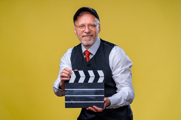 Hombre guapo senior sosteniendo un badajo de cine. Hombre vestido con traje sin chaqueta. Persona aislada contra el fondo amarillo. Produccion de pelicula.