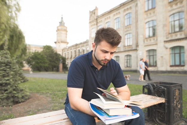 Hombre guapo sentado en un banco en un campus universitario y leyendo un libro.