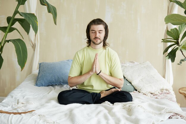 Hombre guapo sentado en la cama en posición de loto y namaste, concepto de atención y yoga, espacio de copia