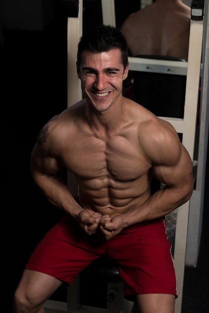 Foto hombre guapo sentado fuerte en el gimnasio y flexionando los músculos muscular culturista atlético modelo de fitness posando después de los ejercicios