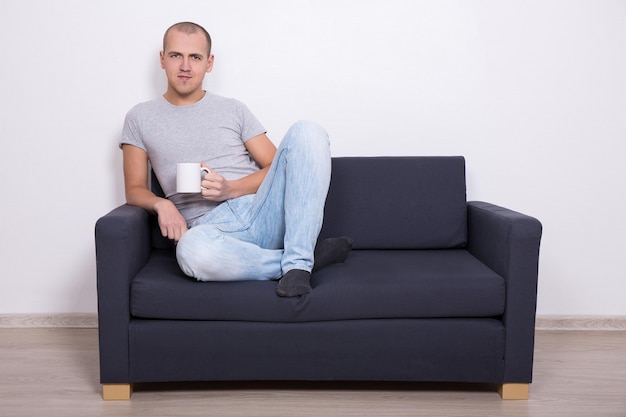 Hombre guapo sentado en el sofá y viendo la televisión con una taza de té o café