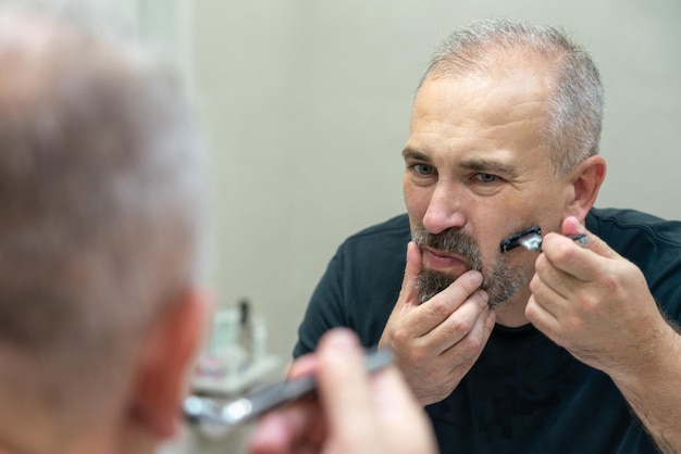 Hombre guapo serio mirando en el espejo y afeitándose los bigotes