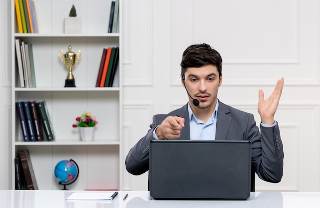 Hombre guapo de servicio al cliente en traje gris con computadora y auriculares agitando la mano y señalando