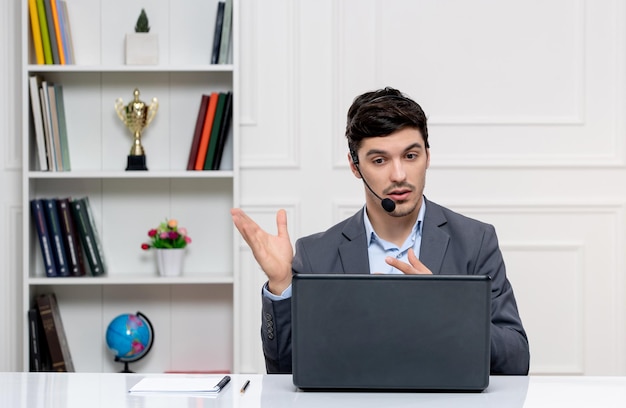 Hombre guapo de servicio al cliente en traje gris con computadora y auriculares hablando por videollamada