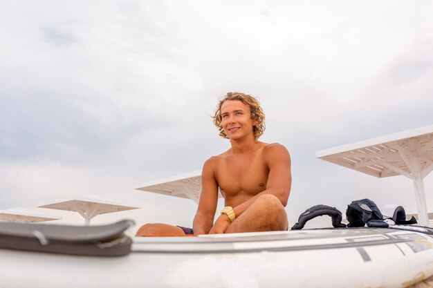 Hombre guapo se sienta en la playa con tabla de surf en blanco blanco esperar a que la ola surfear en la orilla del mar