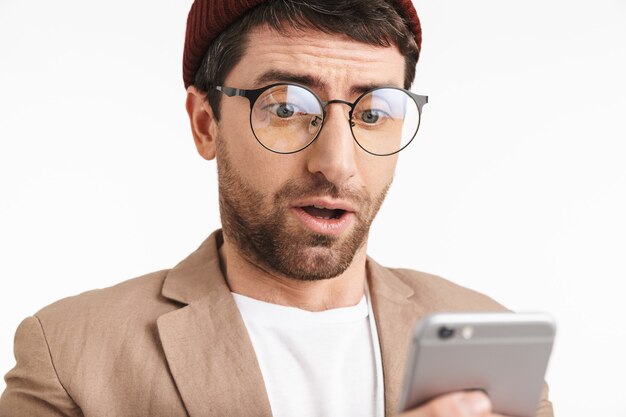 Hombre guapo con sombrero hipster sonriendo mientras sostiene y usa el teléfono inteligente aislado sobre la pared blanca