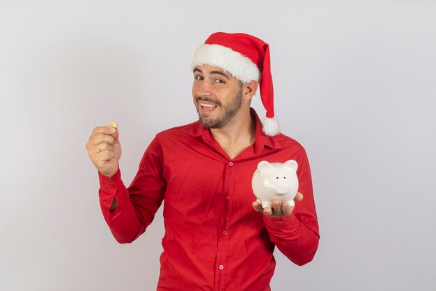 Hombre guapo con sombrero de navidad sosteniendo una alcancía sobre fondo blanco Concepto de ahorro de fin de año