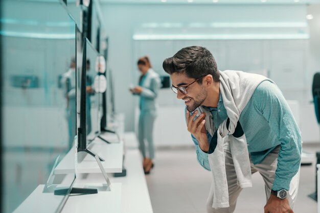 Hombre guapo sonriente con gafas y vestido casual elegante mirando el precio y las especificaciones de la televisión que quiere comprar