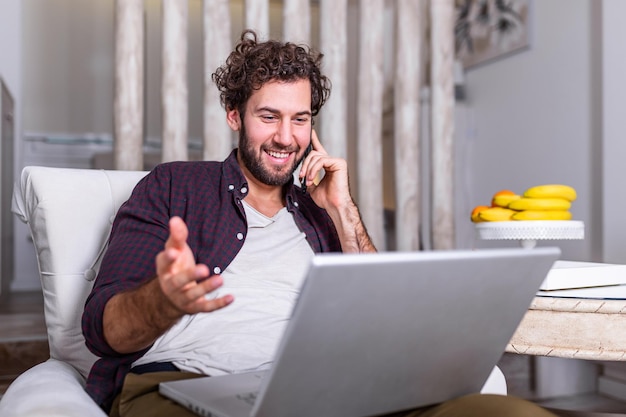 Hombre guapo sonriente hablando por teléfono inteligente y usando la computadora portátil mientras está sentado en casa. Carrera autónoma. Chico hablando por teléfono móvil con ordenador portátil sentado en el suelo en casa. Espacio libre para texto