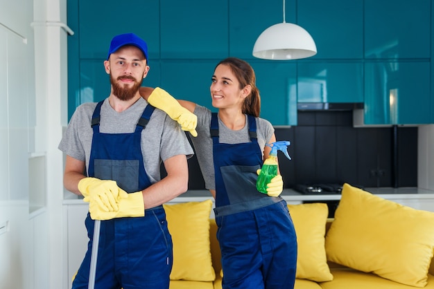 Un hombre guapo sonriente y una joven atractiva como limpiadores profesionales permanecen juntos en la cocina contemporánea con detergentes.