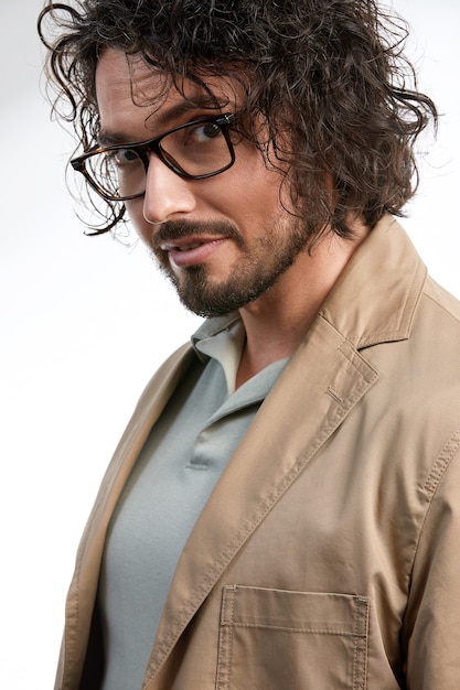 Hombre guapo, sonriente, joven con cabello largo oscuro y gafas elegantes aisladas sobre fondo gris, con ropa elegante, positivo y exitoso en la carrera y los negocios.