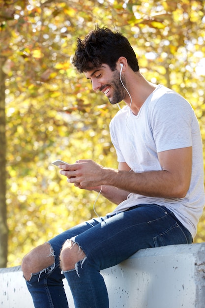 Hombre guapo sonriente sentado afuera con auriculares y teléfono móvil