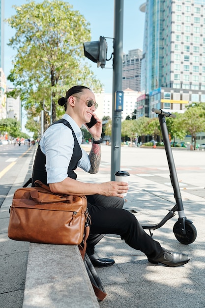 Hombre guapo sonriente sentado en el parapeto, bebiendo para llevar y hablando por teléfono con su colega