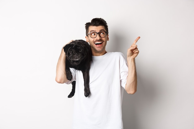 Hombre guapo sorprendido con gafas, sosteniendo lindo perro pug negro en el hombro