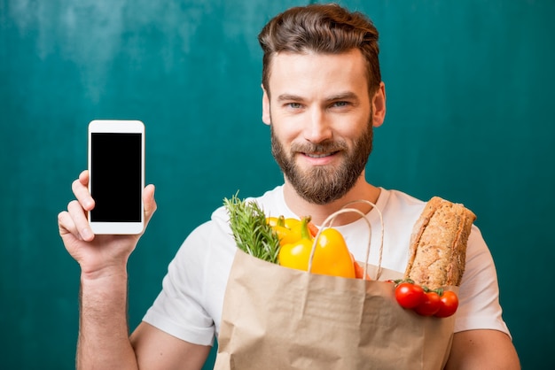 Hombre guapo sosteniendo una bolsa de papel llena de comida sana con teléfono móvil en el fondo verde