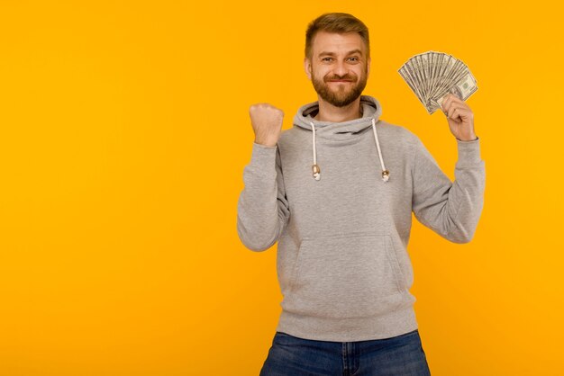 Hombre guapo con una sudadera con capucha gris disfruta de ganar el dinero de la lotería dólares sobre un fondo amarillo - imagen