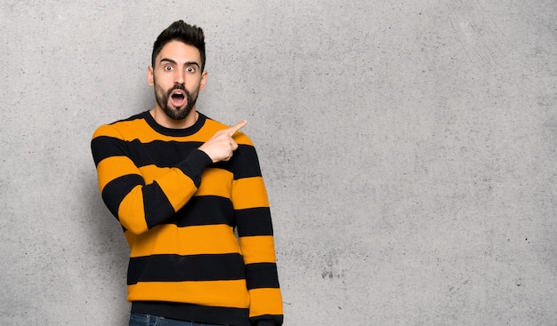 Hombre guapo con suéter rayado sorprendido y señalando el lado sobre la pared con textura