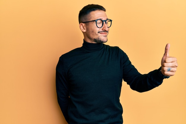 Hombre guapo con tatuajes con suéter de cuello alto y gafas con aspecto orgulloso, sonriendo haciendo un gesto de aprobación hacia un lado