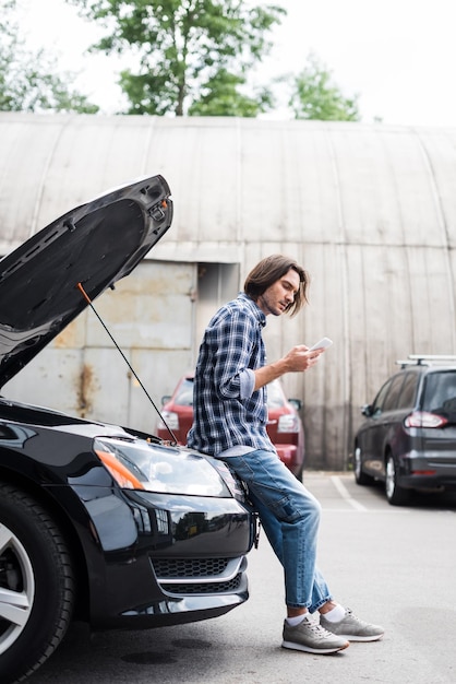 Hombre guapo con teléfono inteligente de pie cerca de auto roto con concepto de seguro de automóvil de maletero abierto