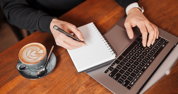 Hombre guapo trabajando en equipo portátil en el lugar de trabajo. Empresario escribiendo información en la computadora en la mesa de trabajo con café y bloc de notas