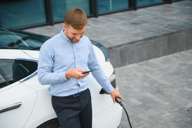 Hombre guapo en traje de negocios navegando por Internet en un teléfono inteligente moderno mientras espera que se cargue un coche eléctrico