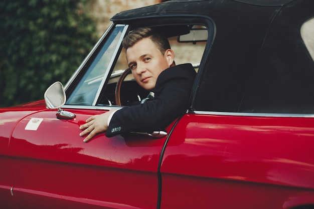 Hombre guapo en traje negro se sienta en un coche italiano retro rojo