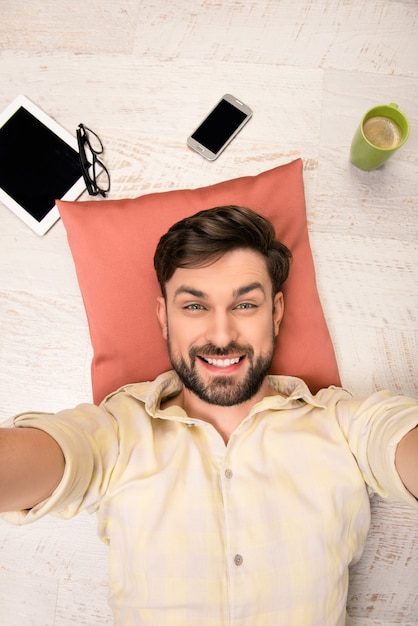 Hombre guapo tumbado en el suelo sobre la almohada y haciendo selfie