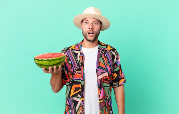 Hombre guapo viajero mirando muy conmocionado o sorprendido y sosteniendo una sandía. concepto de vacaciones