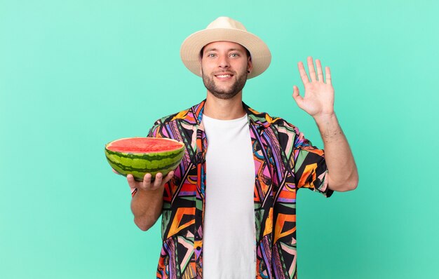 Hombre guapo viajero sonriendo felizmente, saludando con la mano, dándote la bienvenida y saludándote y sosteniendo una sandía. concepto de vacaciones