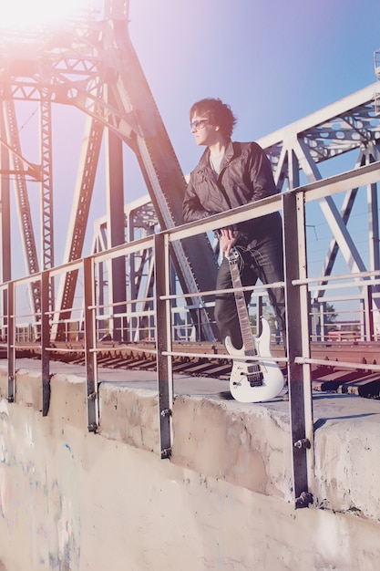 Un hombre con una guitarra eléctrica en el ferrocarril. Un músico con una chaqueta de cuero con una guitarra en la calle de la zona industrial. Guitarrista en el puente.