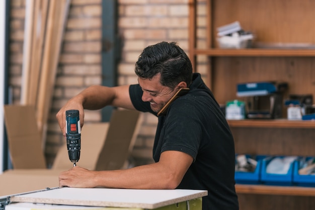 Hombre hablando con un móvil mientras perfora un mueble en un taller