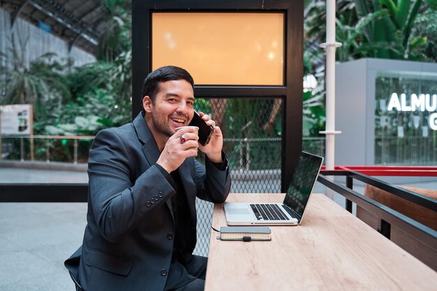 hombre hablando en su teléfono inteligente mientras está sentado en un bar bebiendo café y trabajando en su portátil