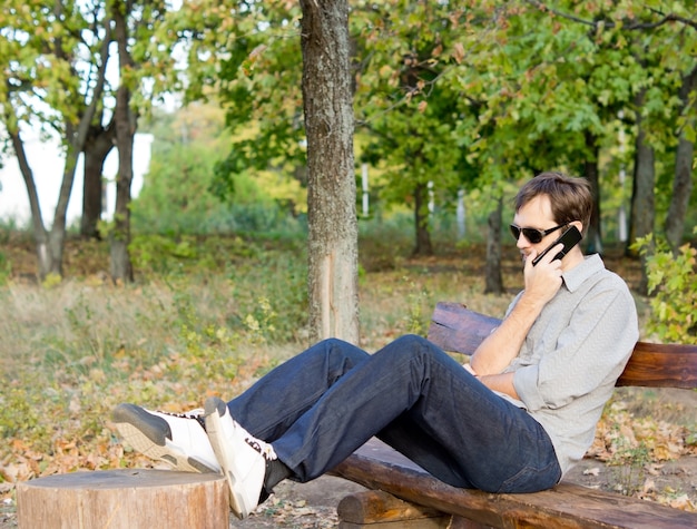 Hombre hablando por su teléfono móvil sentado relajándose en un banco de madera en el campo con los pies sobre el tocón de un árbol