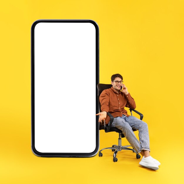 Hombre hablando por teléfono celular cerca de gran smartphone sentado fondo amarillo