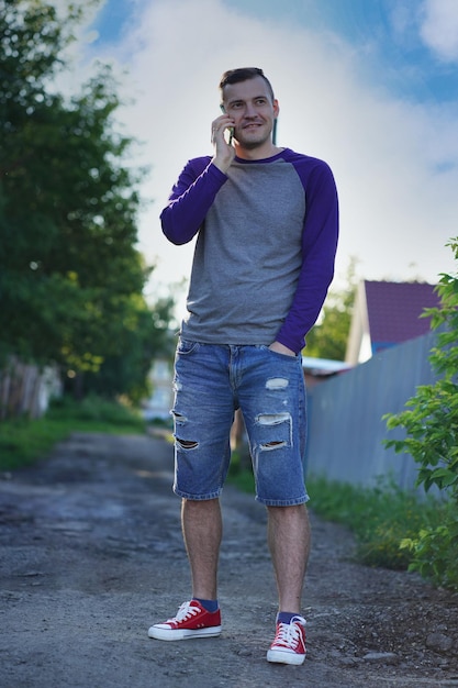 Hombre hablando por teléfono inteligente en un suburbio Hombre adulto con ropa informal respondiendo llamadas telefónicas y mirando hacia otro lado mientras está parado en el fondo borroso de una calle suburbana